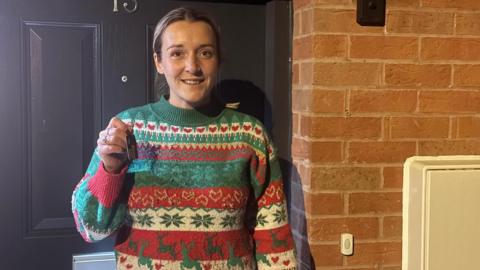 A woman in a Christmas jumper smiling outside a dark grey door. In her right hand she is holding a key, to her right is brickwork and a doorbell illustrating she is standing in front of a house.