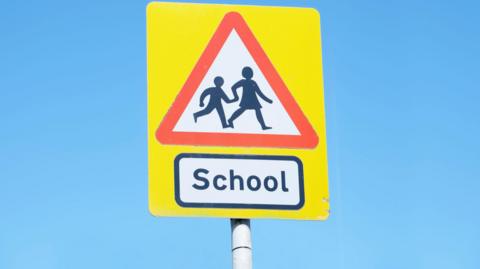 A yellow road sign which has a red triangle on it and a picture of two people holding hands and crossing the road. Underneath the triangle there is a rectangle with the word school written in it in black letters.