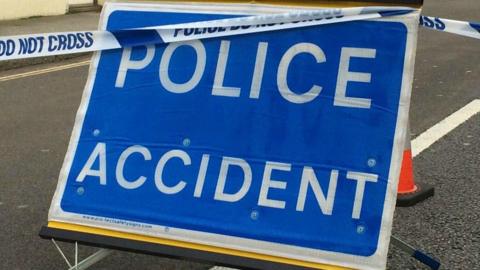 A sign which is blue and has white text reading - top line - POLICE - bottom line - ACCIDENT, is in the middle of a road. Police tape can be seen at the top of the picture.