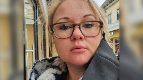 A head and shoulders shot of Gemma Whitehead. She has blonde hair tied back and is wearing brown glasses. She is wearing a grey coat and a black and white scarf. 