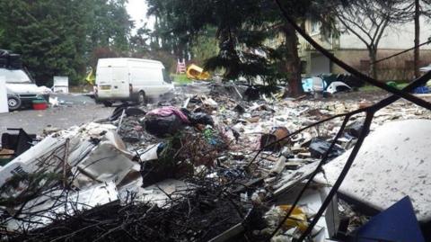 Rubbish, including paper, clothes and white goods, is piled up around knee-high on a tarmac courtyard next to a white building. The area is bordered by tall trees. There are two white vans parked on the courtyard too. 