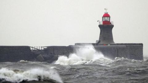 Damage is visible to the pier structure which has large rocks in front of it. There is a lighthouse, large waves and three birds in flight.