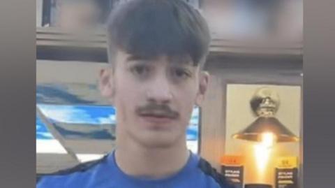 A picture of Taylor wearing a blue shirt with a white Nike logo on it. He has a short moustache and short hair.
