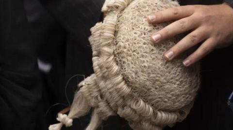 A barrister's hand is seen holding a wigs, which is known as a peruke