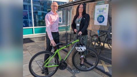 A man in a pink shirt and black trousers is standing next to a woman in a suit jacket and multi-coloured dress. They are standing in front of a green bike near a bike rack.