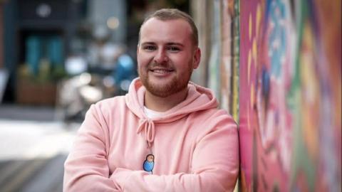 Gary Harper has short light brown hair and a short beard. He is wearing a pink hooded top and is smiling at the camera. He is leaning against a painted wall.