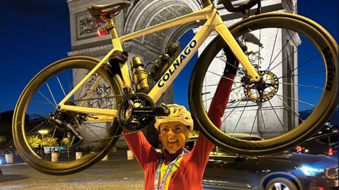 A smiling Lyndsey Blair, raises her yellow bike in triumph after crossing the finish line in Paris. She is wearing black cycle shorts, a red long-sleeved cycle top and a yellow helmet. She has a medal round her neck
