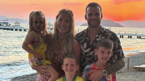 The family pose for a family photograph on a sunny beach. Glen and Gemma smile at the camera along with their daughter and two sons. The sea and a pier can be seen in the background.
