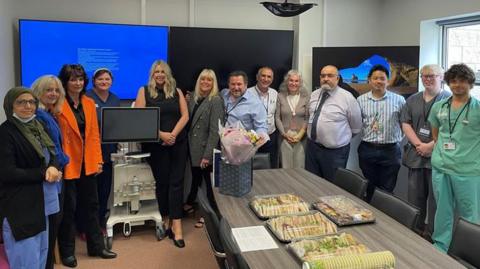 Doctors and medical staff line up next to the machine which has a widescreen on top and a cream body. Julie Stokes from Mannin Cancers Support Group stands in the middle of the group of thirteen people. They are standing in a committee room with a long table in the middle.