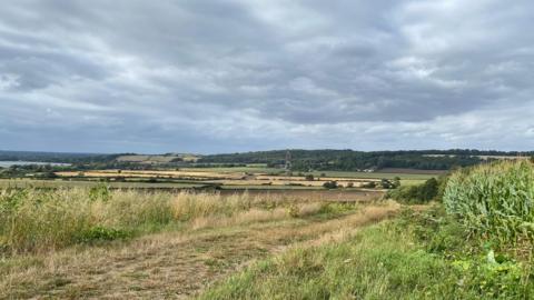 Rolling green grass hills with a dark grey stormy sky overhead. The foreground shows a grass path with a hedge and in the distance you can see electricity pylons, trees and fields. 