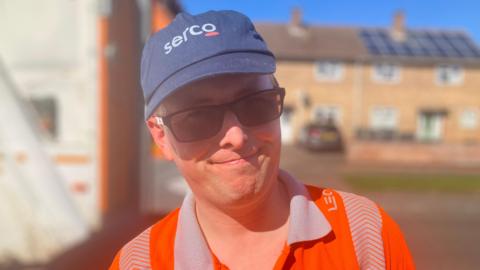 Robert Butler wearing a blue cap, sunglasses, and high-vis jacket on his bin round. 