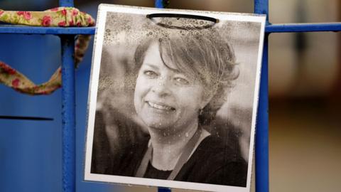 A printed black and white photograph of Ruth Perry fixed to a blue-coloured fence. Mrs Perry is smiling in the photo. 