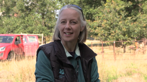 Jane Birch, a woman sitting outside in parkland. there is a red pickup truck behind her. She is wearing a green fleece and dark grey National Trust branded gilet. She has light-coloured hair and a pair of sunglasses on her head. The photo was taken on a sunny day.