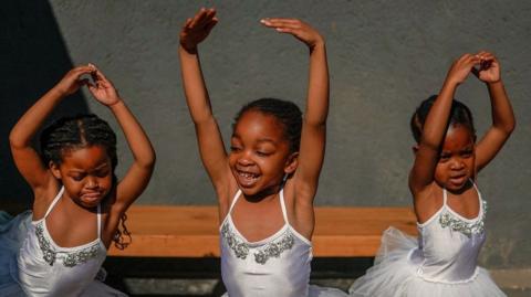 Three girls wearing white leotards and tutus stretch their arms out.