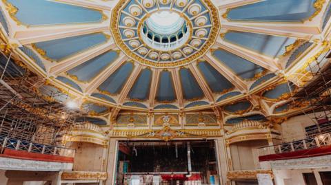 The image shows the interior of a theatre, with high ceilings where blue and gold can be seen; scaffolding can also be seen on the balconies around the building.