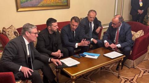 Starmer, Macron, Zelensky, Tusk and Merz in formalwear and sit on sofas around a table, on which sits a phone and papers. 