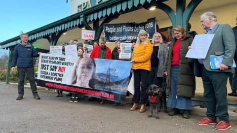 Protestors are gathered out Strathpeffer Pavilion. They are holding banners and have a dog with them.