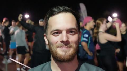 Nathan Parris is pictured smiling to camera. He is pictured at night on an athletics track with numerous other runners behind him. He has dark hair, a beard and is wearing a grey top.