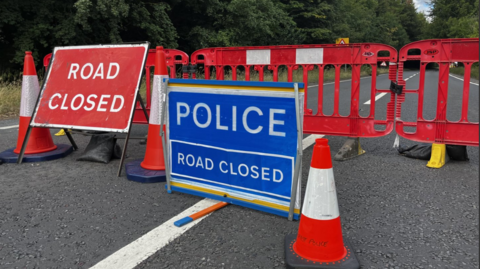 A blue and white police road closure sign stands next to a red and white one with traffic cones in between and red plastic barriers behind on a tree-lined road