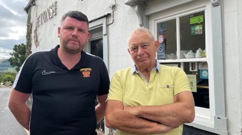 Josh (left) and Tim (right) pictured outside the pub. Josh is wearing a black sports tshirt and Tim is wearing a yellow polo shirt. Josh has his hands behind his back and Tim has his arms folded - they both wear serious expressions. The pub behind them has peeling, cracked white plaster on the outside, and the first three letters have fallen off the sign, so it just reads 'E TUNS INN.'