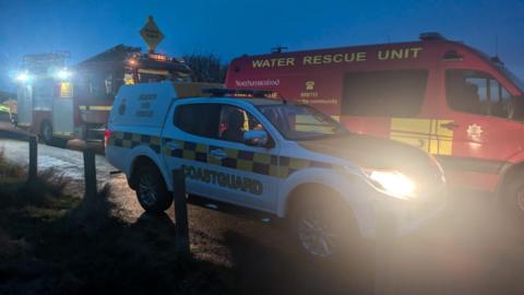 Coastguard and fire service vehicles parked on a narrow road in the dim light. 