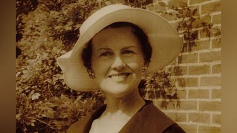 A headshot photo of Muriel McKay standing outdoors with a wall behind her that is partially covered in the creeper wisteria. She wears a flattering white rimmed hat, cluster earrings and a dark coloured coat. The photo is sepia coloured.