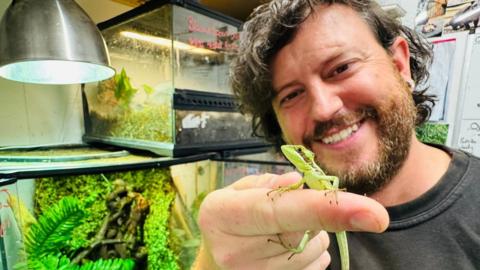 A man with brown curly hair and brown facial hair is holding his finger up to the camera. Sitting on his finger is a small green iguana. Behind the man are glass tanks with green foliage in them