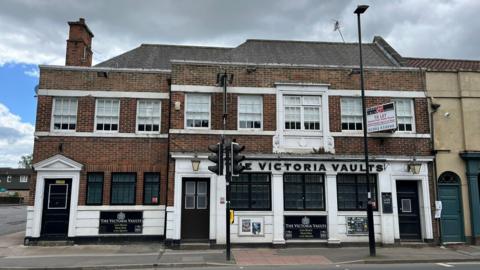 A large brick building is shown across a road. There are traffic lights and a streetlight in the foreground, and the lower half of the building is painted white. The sign says 'The Victoria Vaults' and has large windows. There's three doors to the building, and it has 14 windows in total. There are aerials on the roof and a chimney, as well as a dark green door to the right of the photo which is part of a connecting building. 