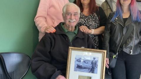 Stuart MacDonald smiling at the camera wearing a black work fleece and a green shirt, and holding a frame with an old photo of him on it in a suit standing next to a lake