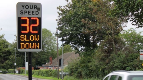 A speedometer that says "Your speed, 32, slow down" as a silver car, that is partly visible, is driving through a residential area. 