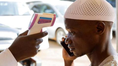 A man dressed in white is seen on the right of the image on his mobile phone as his temperature is taken with a contact-free temperature gun. A number of cars are out of focus in the background of the picture.