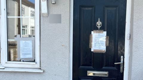A black front door has an A4 sheet of paper full of text sellotaped to the middle of it. There is a similar notice on the window to the left of the door.