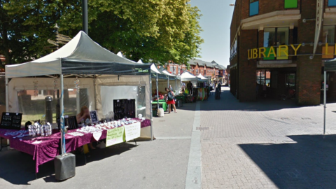 A street with several white market stalls on the left hand side selling products. On the right is a large building with a yellow sign that reads "library". There are green trees behind the market stalls.