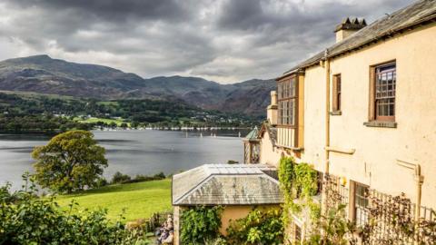 Brantwood - a substantial, pink-hued house stands beside Coniston Water. In the distance, hills and mountains rise above the village.