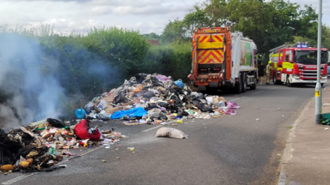 A large pile of discarded waste spread across a road, with smoke rising from a section of the rubbish in the foreground. A refuse collection vehicle and a fire engine are positioned nearby. Dense hedgerows line one side of the road, while a pavement and streetlight are visible on the opposite side.