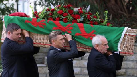 Hefin David's coffin draped in a Welsh flag with red roses on the top, being carried by six people, with three men seen from the side.