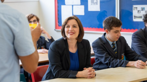 Phillipson wears a black suit jacket over a royal blue dress with a silver necklace. Her hair is worn in a dark brown, chin-length bob. She wears bright red lipstick and is smiling at a teacher, who can partially be seen in the foreground on the left. There are pupils sitting around her, all wearing a navy school uniform with a pastel blue shirt and a navy tie with pale blue and gold diagonal stripes. 
