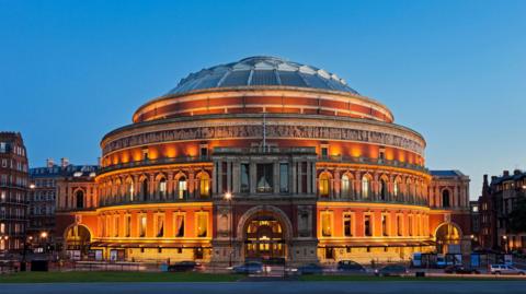 Exterior view of the Royal Albert Hall in London at dusk, with its red brick facade illuminated by golden lighting against a clear blue sky.