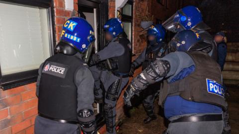 Five police officers in helmets and stab vests step through a door they have just knocked in