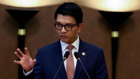 A man wearing black-rimmed glasses and in a blue suit with a red tie speaking into two microphones, holding up his hand as he speaks