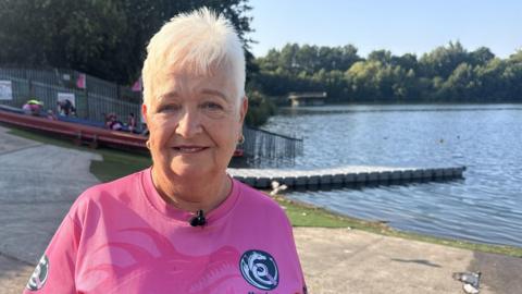 Ann Johnson, who is wearing a pink T-shirt with a dragon logo representing Manchester Dragoneers, is standing in front of a reservoir and smiling at the camera