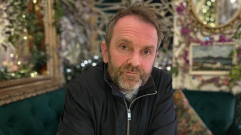 Eamon McCusker in his AMPM restaurant.  He has short, fair hair, blue eyes and a greying beard.  He is wearing a black zipped jacket. 