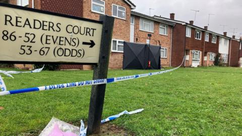 Police tape at the block of flats in Readers Court. In the foreground is a road sign that directs people to Readers Court. The police tape then leads you to the flats. In front of one is a black screen.