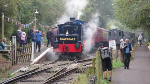 A view of a red and black locomotive on train tracks. There is steam coming from the bottom and sides of the train. On the sides, there are people walking, some taking photos. There is bunting on one platform.