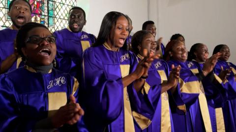 Aberdeen Gospel Choir singing and clapping their hands, wearing purple and gold robes and standing in front of a stained glass window