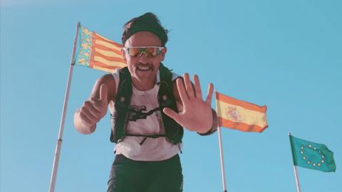 Andy Hobson standing in front of Spanish and EU flags dressed in running gear. He holds up six fingers to symbolise six runs. Andy is wearing sunglasses, has a moustache and a dark headband on. 