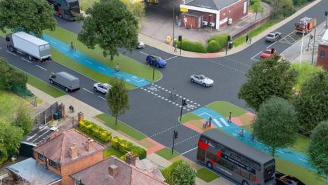 A wide road with a central green area with trees and a cycle path in blue in the centre of it. The traffic on either side of the middle area includes a grey bus, white lorry and several cars.