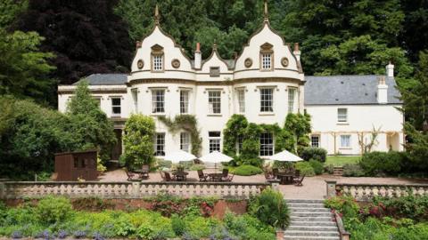 A large 19th century country house nestled amongst trees and bushes. The house is cream coloured and has large windows.