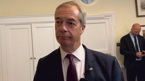 A man looking forwards and to the side of the camera. He is wearing a dark suit and dark purple tie and white shirt. Behind him are white cupboards and another man who is wearing a suit is looking down. 