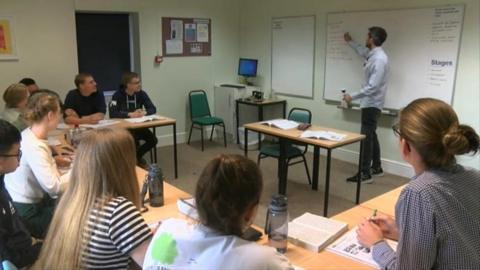 A group of international students sit in a classroom during a lesson as a teacher points at a whiteboard 